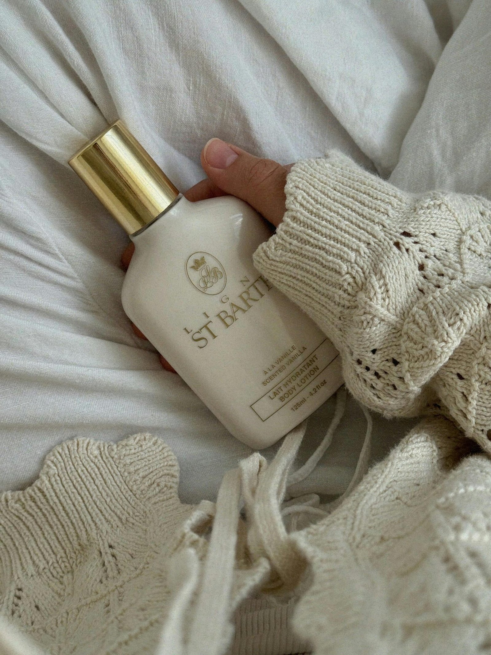 A hand in a sweater holds a luxury body lotion bottle on a white background.